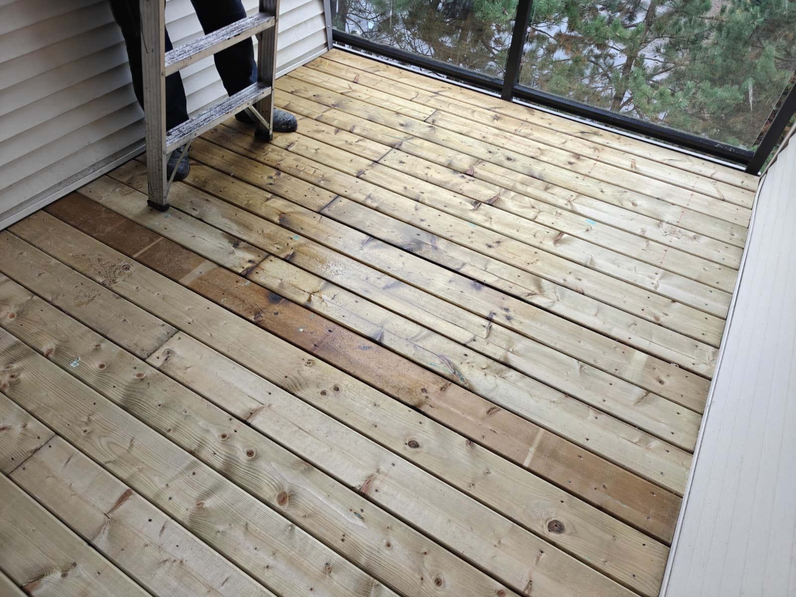 A wooden deck under construction on a porch with a ladder and partially installed boards. A person is standing nearby, wearing black pants and shoes.