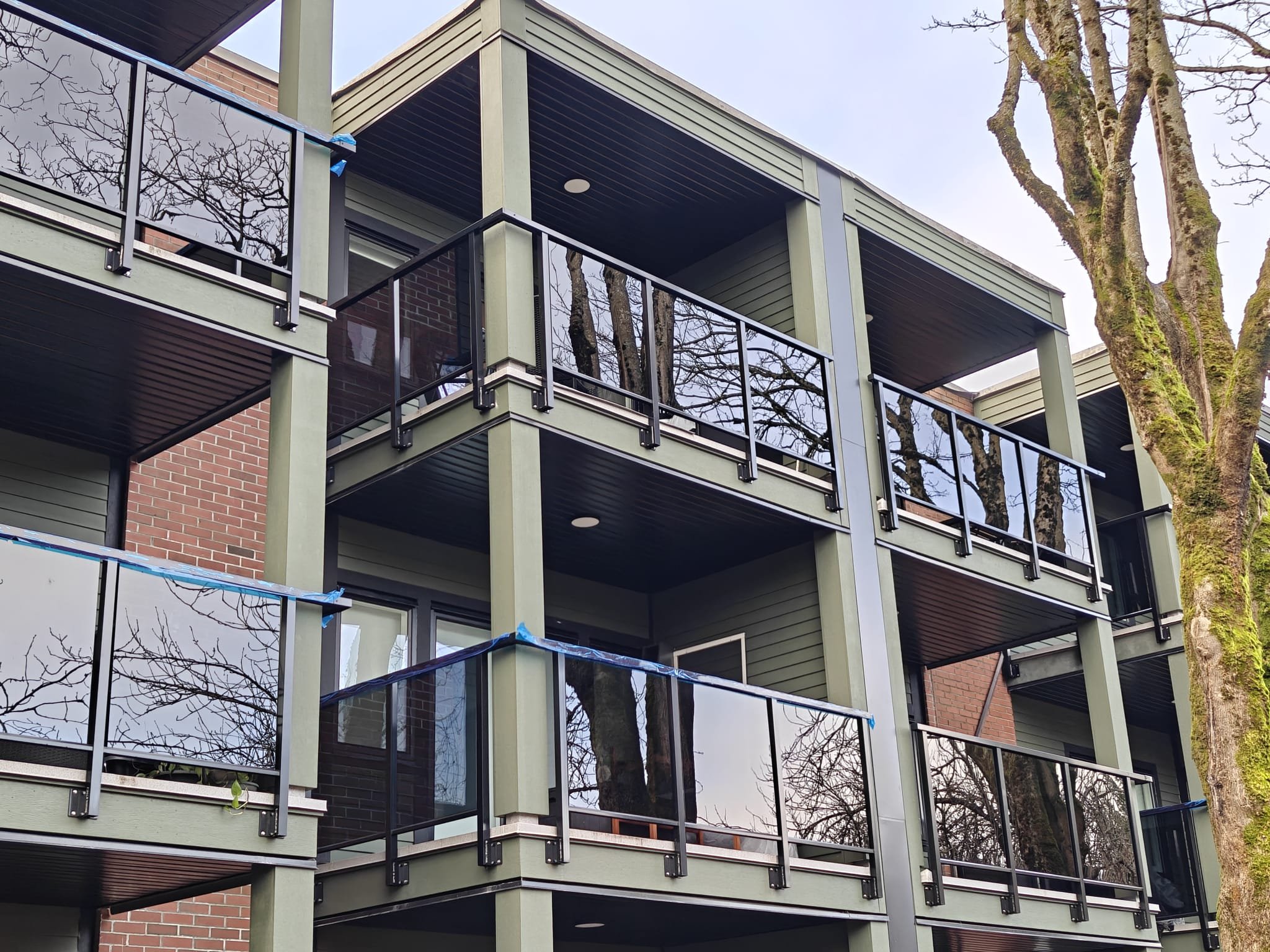 Construction scaffolding around a multi-story residential building with red brick and brick panels, adjacent to greenery and trees, with sidewalk and orange safety fencing along the street.