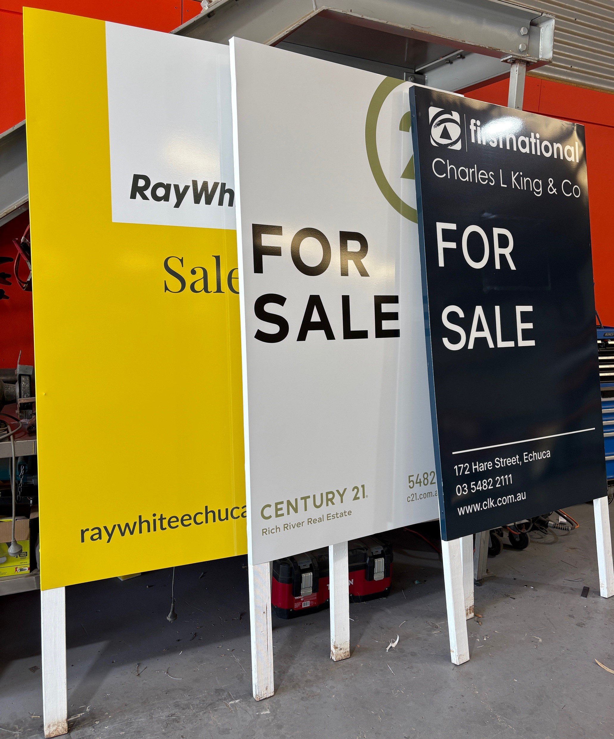 Three sale signs in front of a workshop, advertising properties for sale. The signs are from RayWhite, Century 21, and Charles L King & Co, with the RayWhite sign on the left, the Century 21 sign in the middle, and the Charles L King & Co sign on the right.