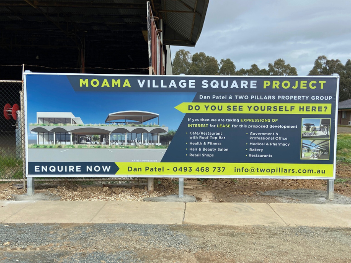 Sign advertising the MOAMA Village Square Project by Dan Patel & Two Pillars Property Group. It features a digital illustration of the proposed development, including cafes, health and beauty salons, retail shops, and government offices. Contact information for Dan Patel and a call to inquire now are also included.