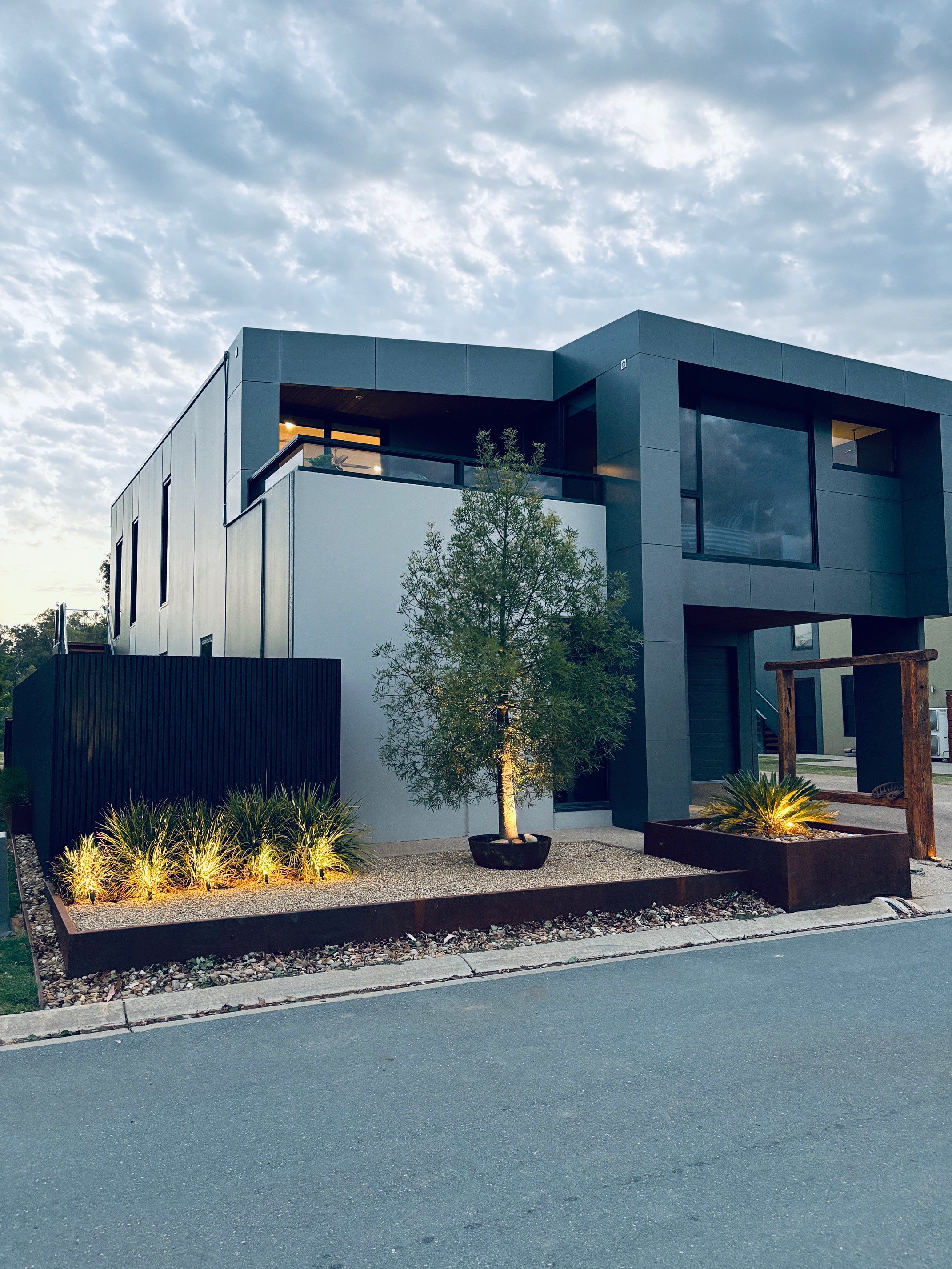 Modern two-story house with a minimalist design, gray facade, and large windows, featuring landscaped front yard with small trees and illuminated plants, under a cloudy sky.