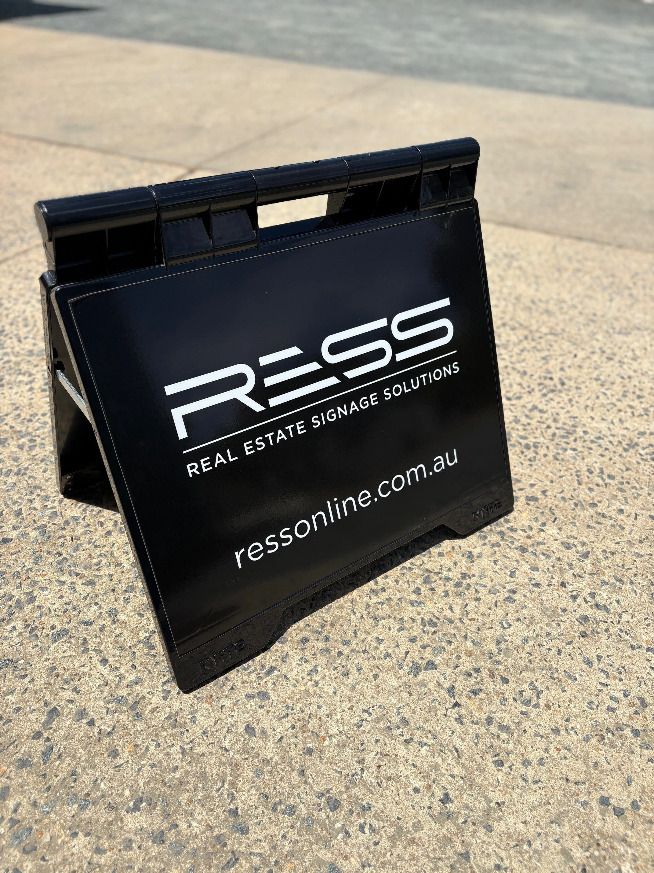 Black A-frame sign with white text advertising RESS Real Estate Signage Solutions and website ressonline.com.au, placed on concrete pavement outdoors.