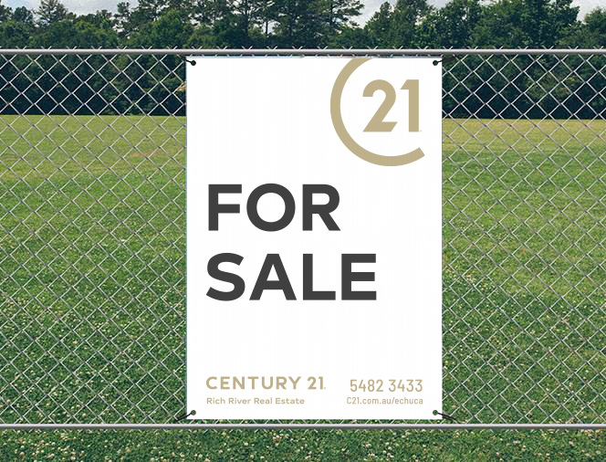 A large vertical banner hanging on a chain-link fence with a grassy field and trees in the background. The banner features the Century 21 logo, the words "FOR SALE" in bold black letters, and contact information for a real estate agent.