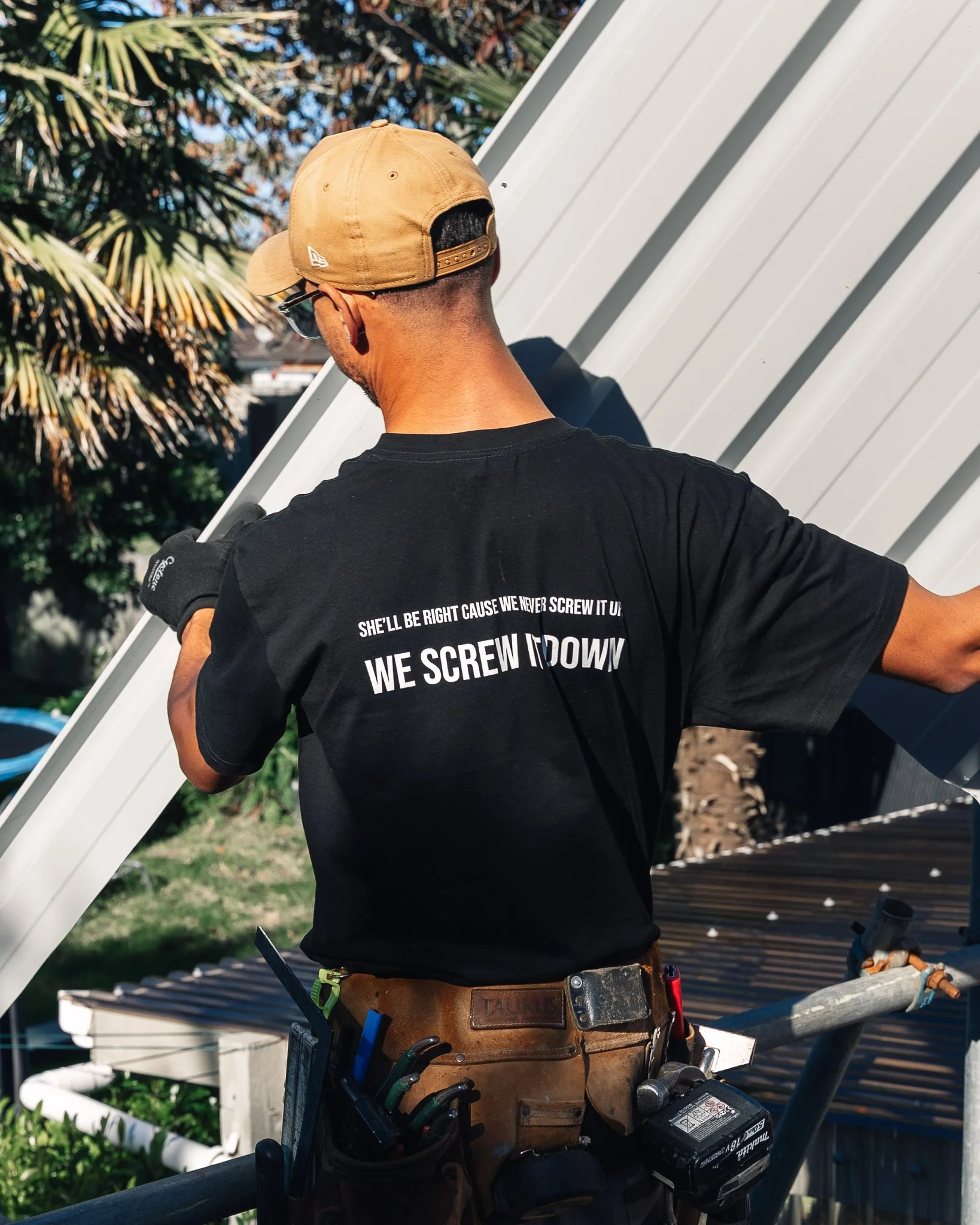 A man in a tan cap and black t-shirt working on a metal structure with tools attached to his belt.