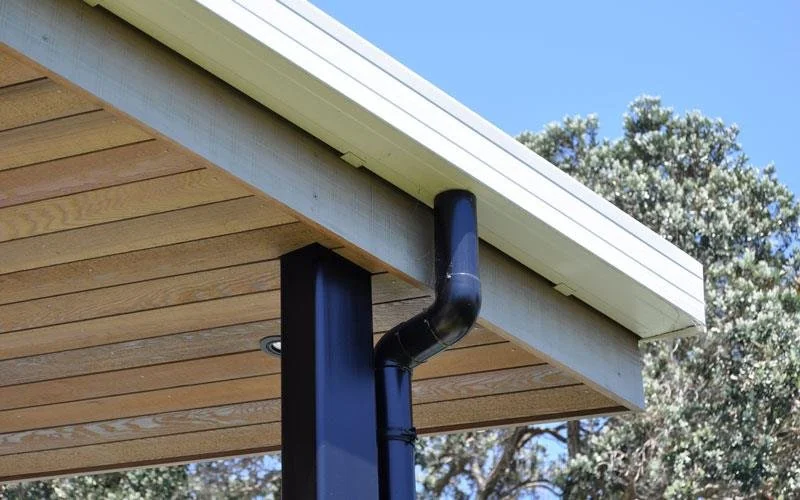Close-up of a house's roofline showing wooden soffit, black gutter downspout, and part of the gutter at the roof's edge with trees and clear blue sky in the background.
