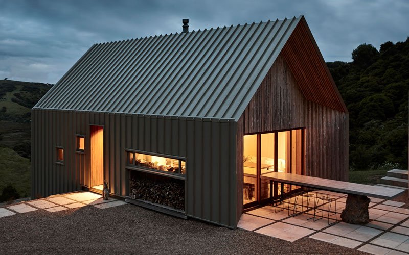 Modern house with metal and wood exterior, large glass doors, and a patio with outdoor furniture, set in a hilly landscape during dusk.