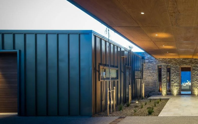 Exterior view of a modern building with a combination of blue metal siding, a stone wall, and a wooden ceiling overhang with recessed lighting.