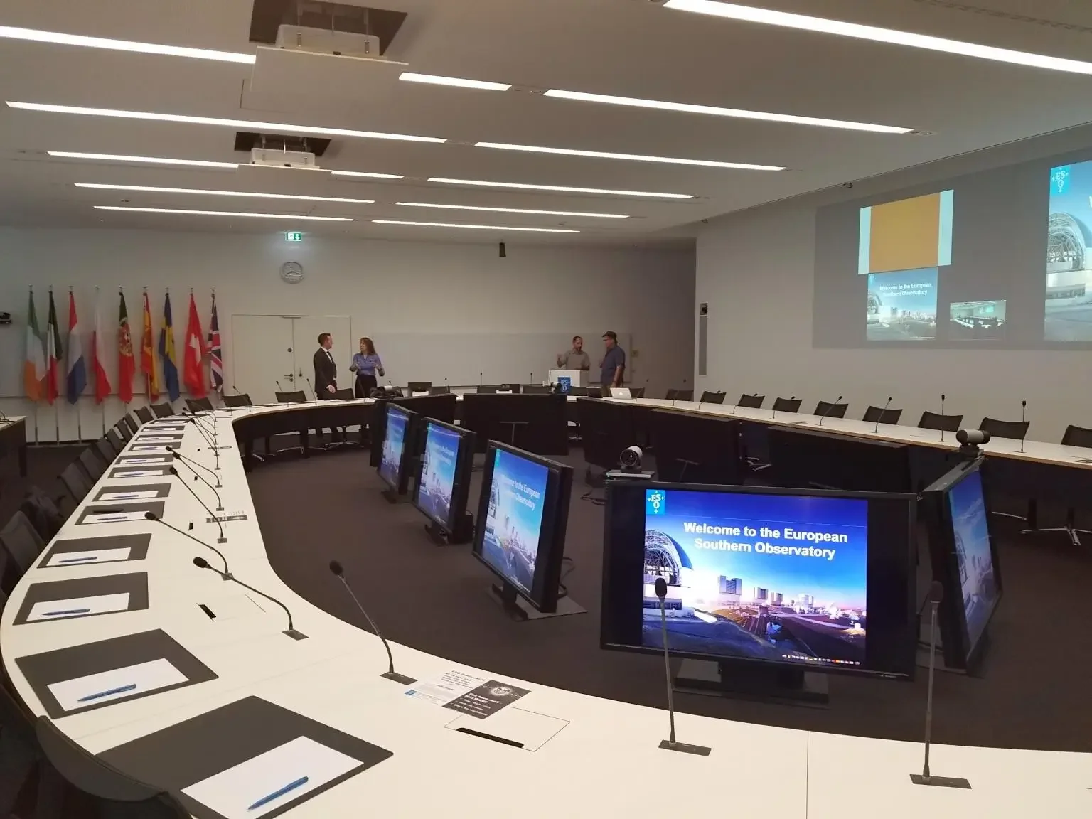 Conference room with a large, U-shaped table equipped with individual monitor screens and microphones, and a projector screen displaying a welcome message for the European Southern Observatory. Five people are standing and talking in the background, and multiple national flags are aligned along the wall.
