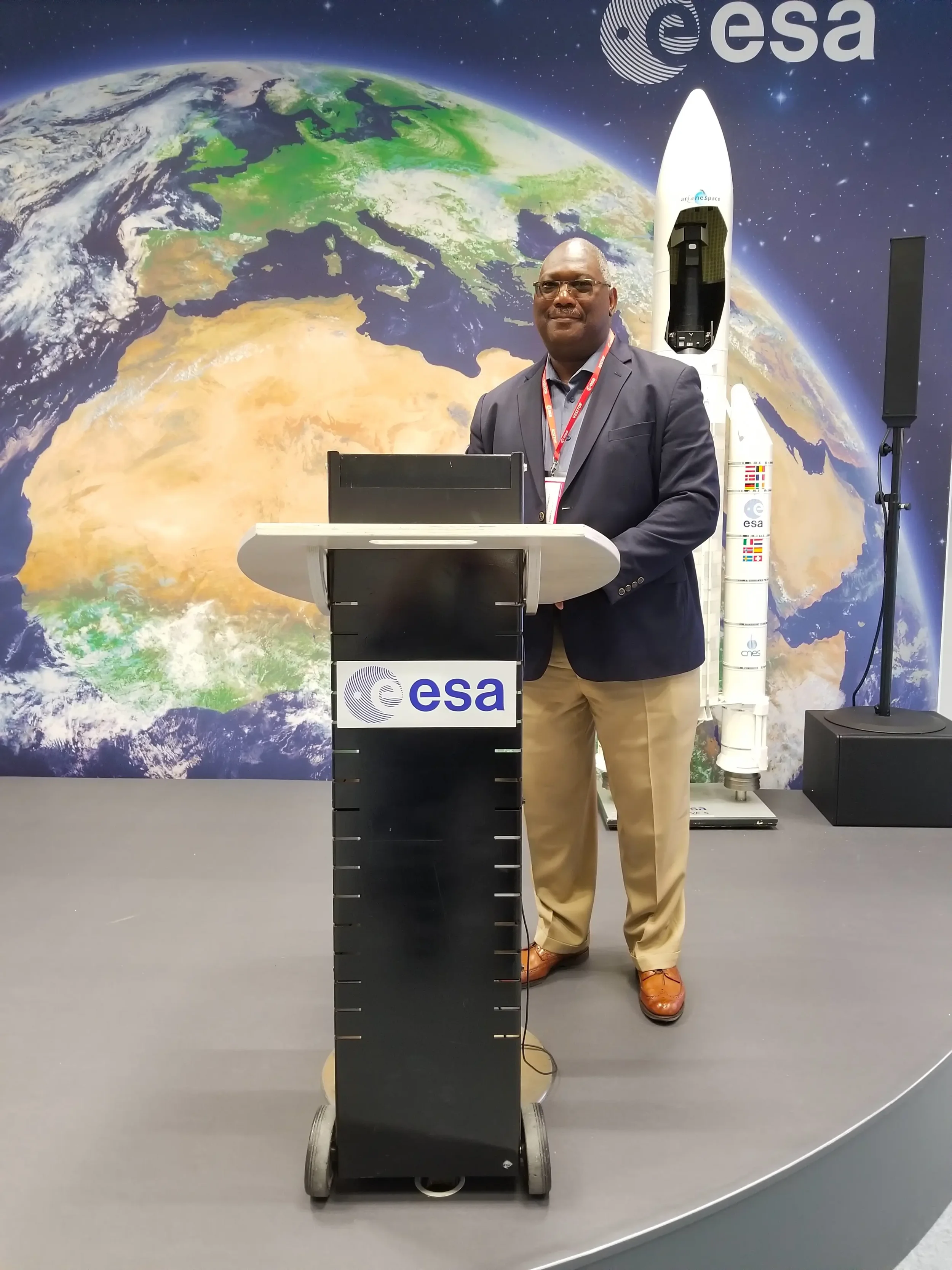 Planetary defense expert L.A. Lewis in a suit, standing at a podium with an ESA logo, in front of a backdrop showing Earth from space and a rocket with ESA branding.