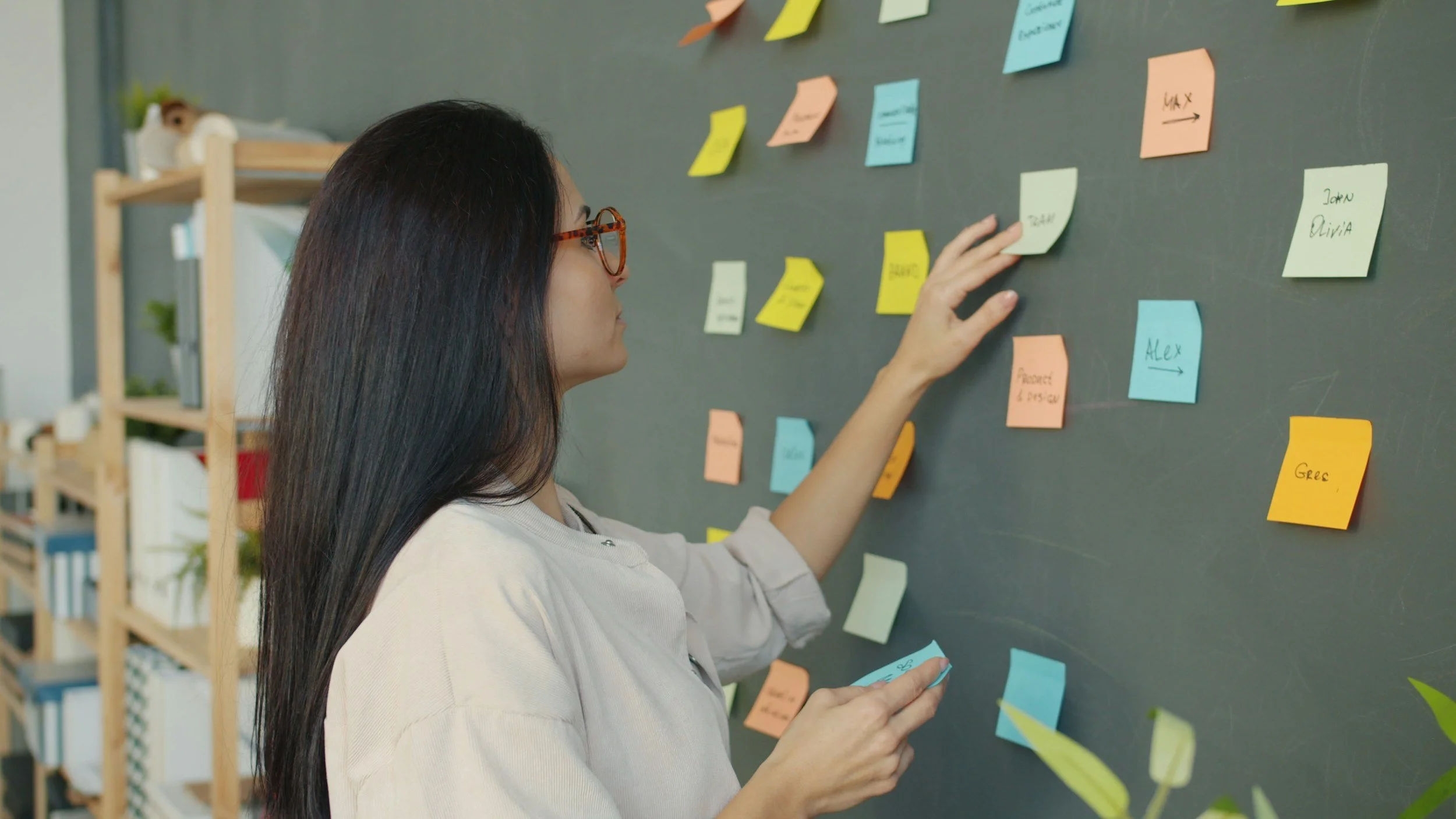 Woman organizing colorful sticky notes on a blackboard in a workspace.