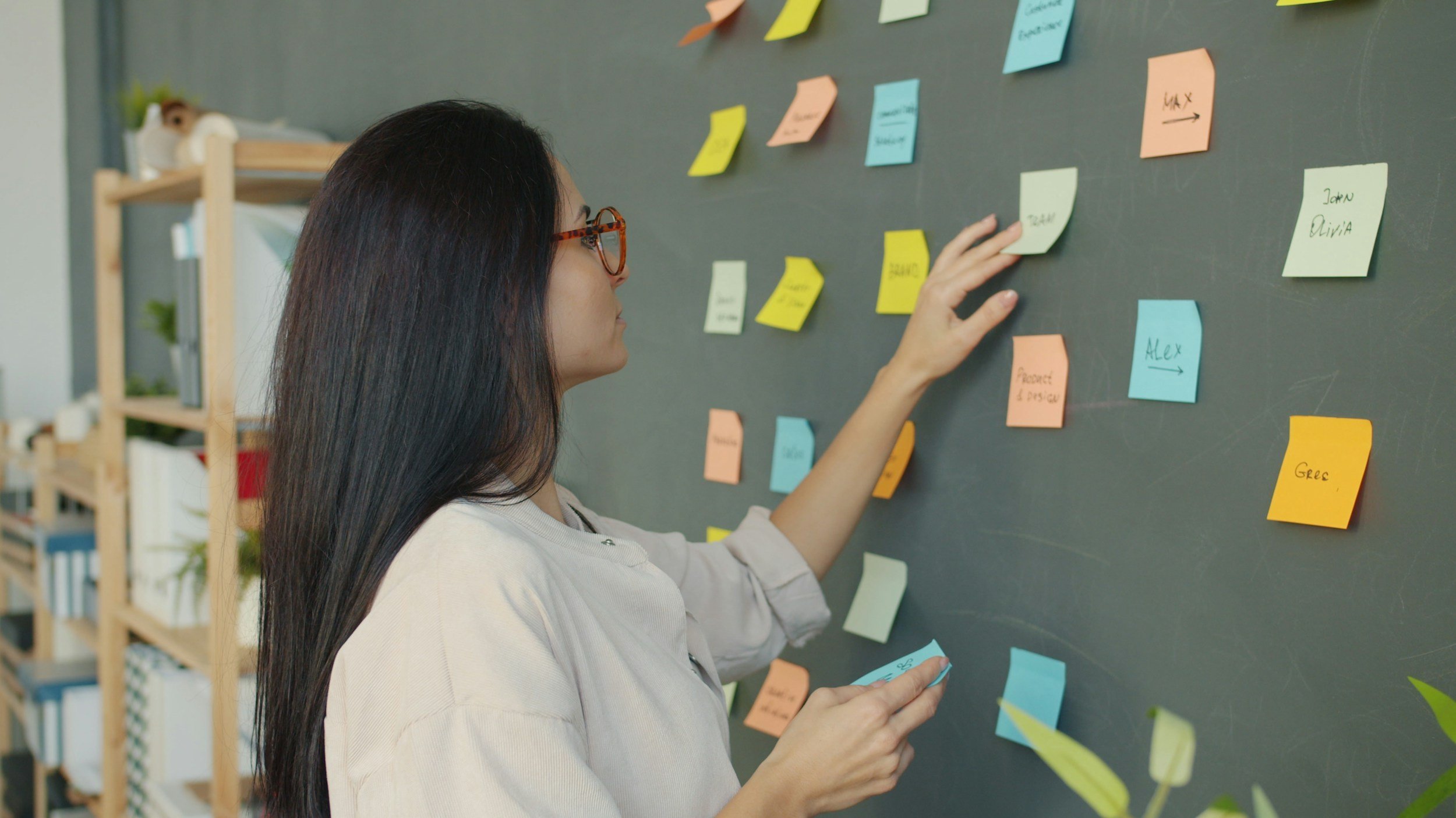 Woman placing sticky notes on a blackboard filled with colorful sticky notes in an office or classroom setting.