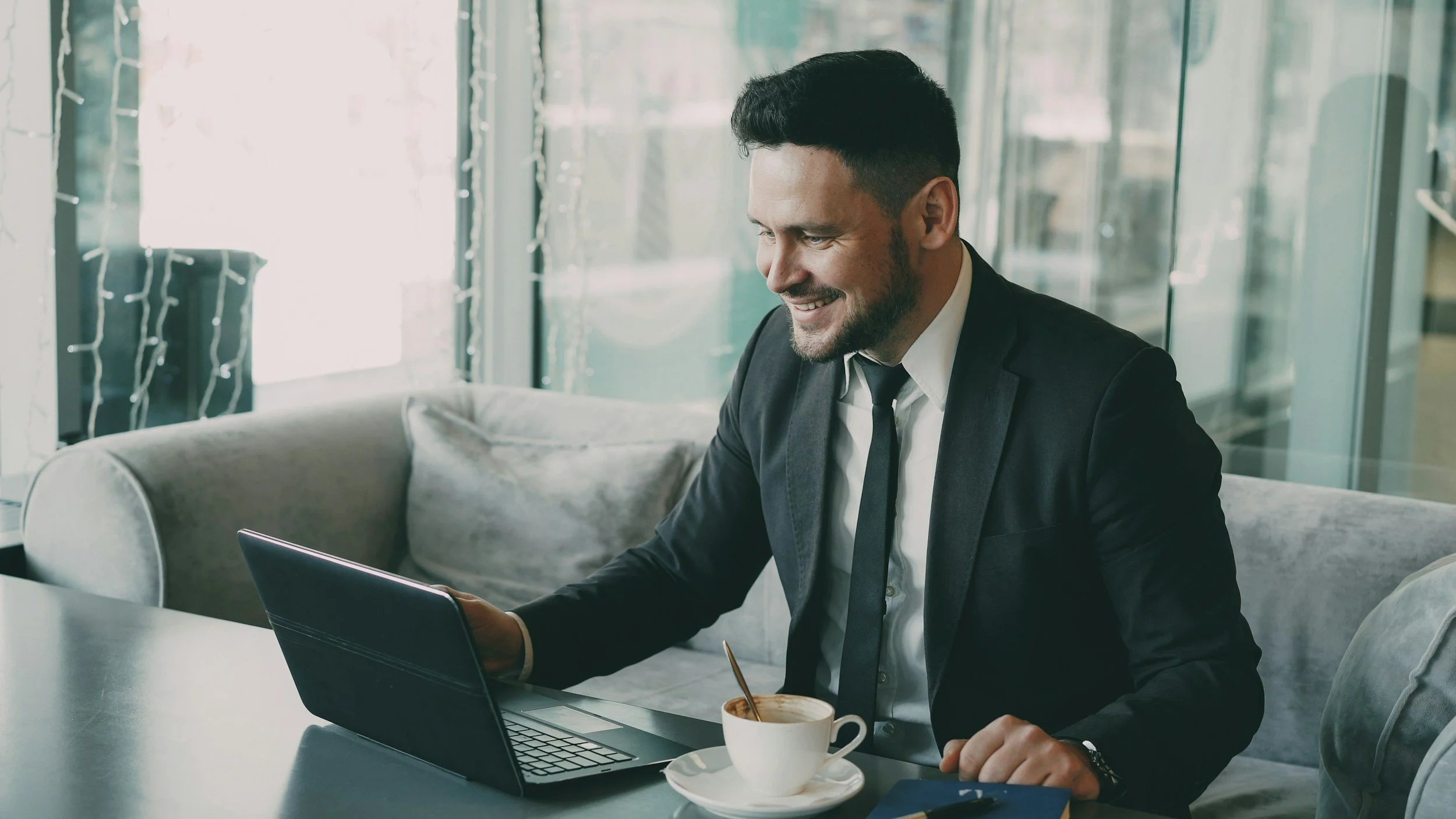 A smiling man in a black suit and white shirt sitting at a table with a laptop and a cup of coffee in a modern indoor setting.