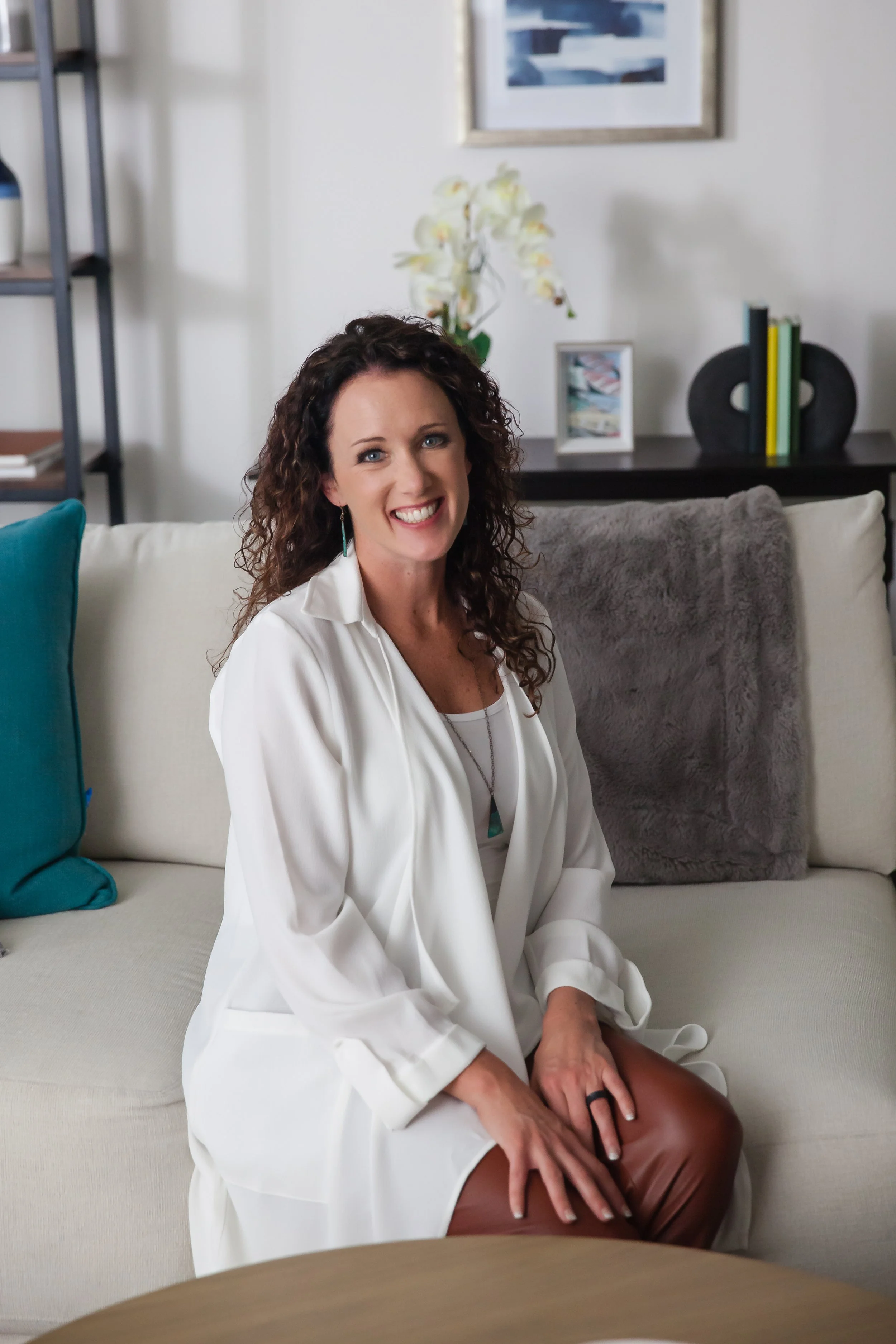 A woman with curly brown hair, wearing a white coat and a turquoise necklace, sitting on a beige sofa smiling at the camera with a living room background including a bookshelf, framed pictures, and an orchid.
