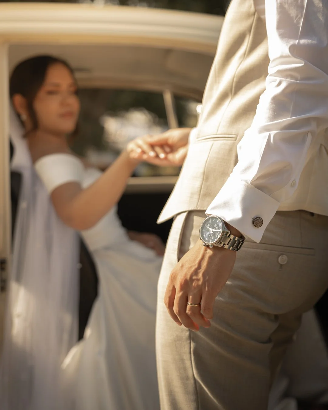 Close-up de um homem com terno bege, usando um relógio de pulso de metal, segurando a mão de uma mulher. A mulher, em movimento dentro de um veículo antigo, usa vestido branco.