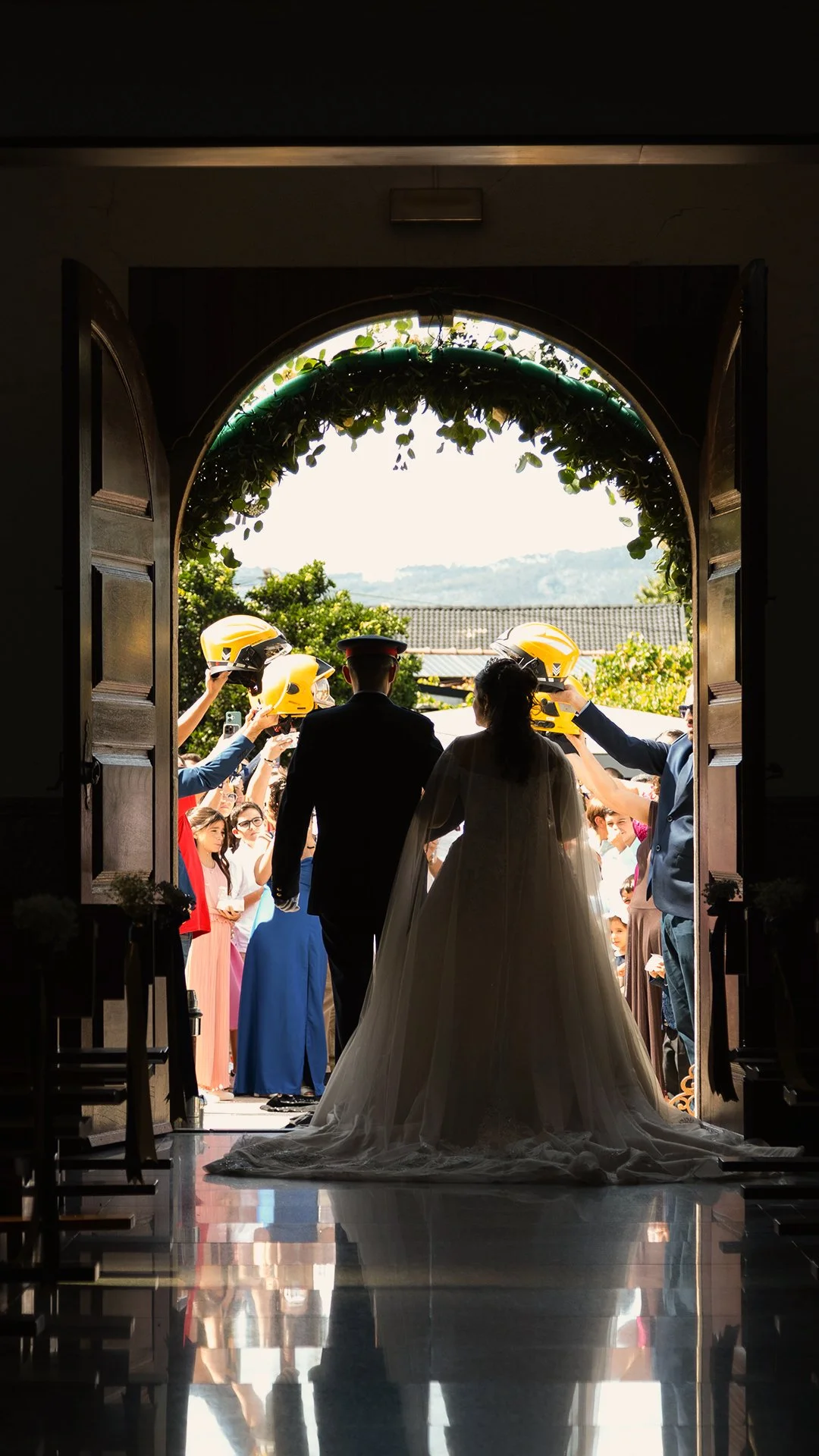 Casal de noivos entrando na igreja, rodeados de convidados e com capacetes amarelos sendo entregues pelos convidados ao ar livre.