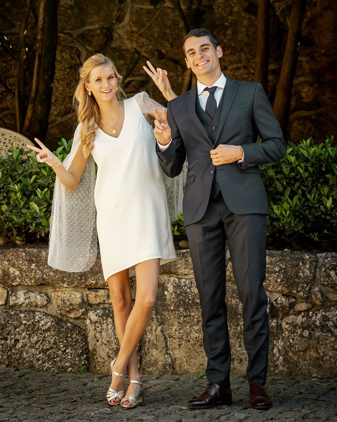 Casal sorridente faz sinais de paz com as mãos, em ambiente ao ar livre com vegetação ao fundo.