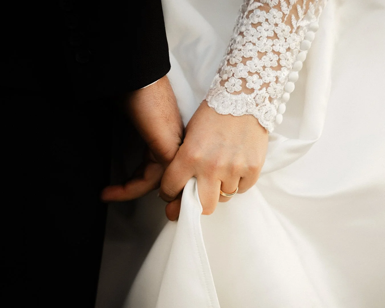 Casal de mãos dadas em casamento, com mulher usando vestido de renda branca e homem de terno preto.