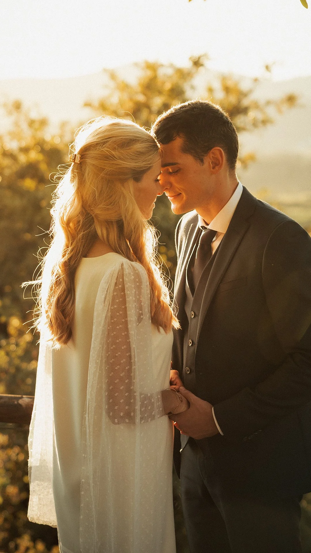 Casal de noivos se olhando com os rostos próximos, segurando as mãos, ao pôr do sol, com árvores ao fundo.