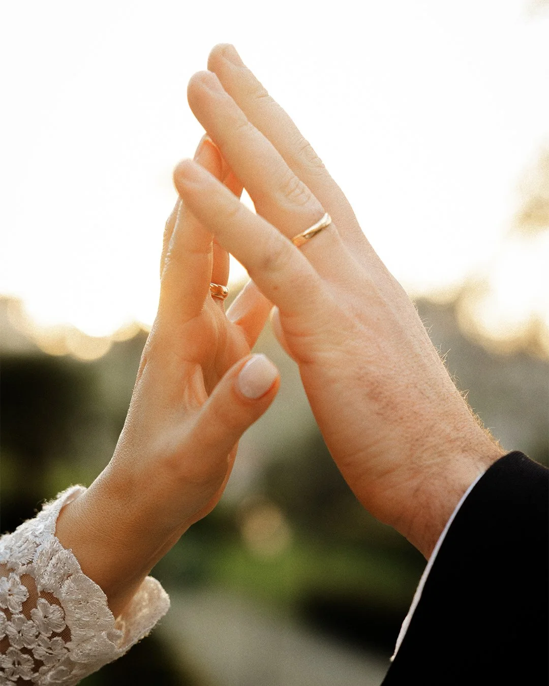 Mãos de um casal se tocando, com uma delas usando um anel de casamento, simbolizando união e compromisso, com fundo desfocado ao pôr do sol.