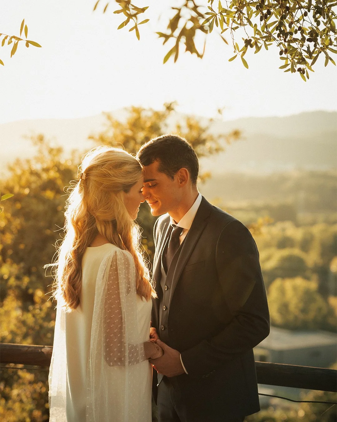 Casal romântico de noivos com cabelo loiro e escuro, se beijando ao pôr do sol em ambiente ao ar livre com árvores e horizonte ao fundo.