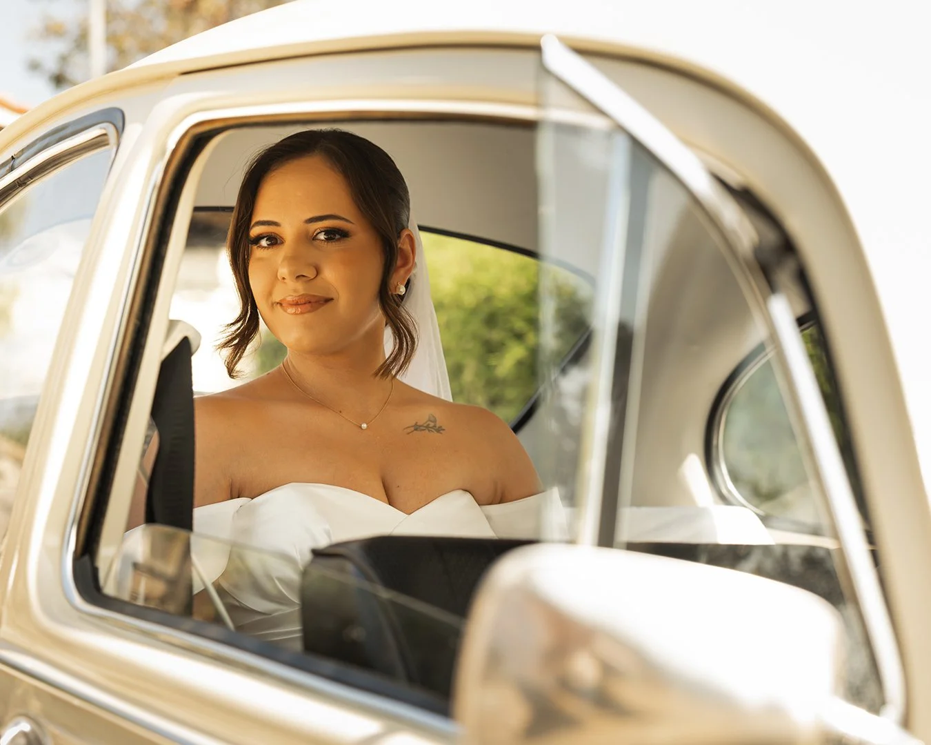 Noiva sorridente olhando pela janela de um carro clássico, vestida de branco e com cabelo de comprimento médio.