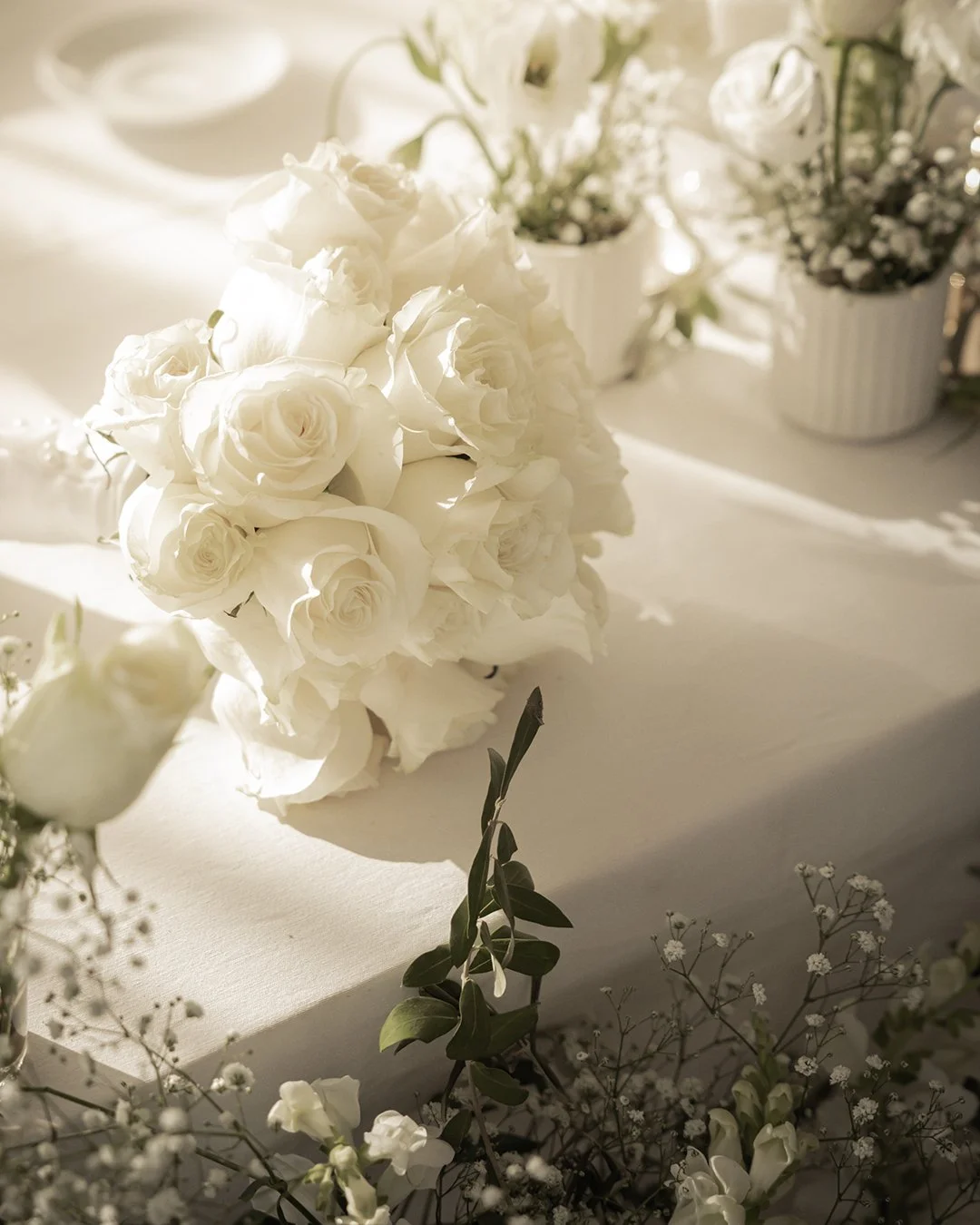 Arranjo de flores brancas, incluindo rosas e gérberas, decorando uma mesa com luz natural.