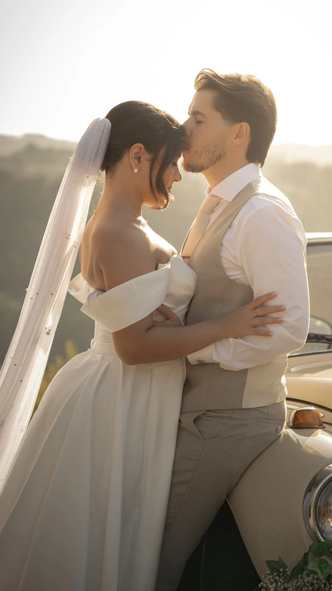 Casal de noivos em um casamento ao ar livre, com o homem beijando a testa da mulher.
