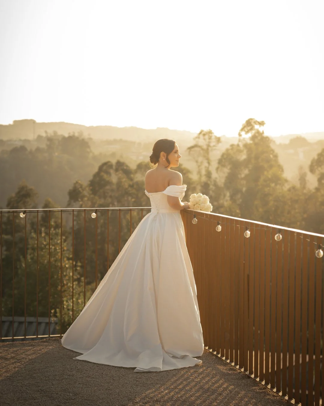 Noivos de vestido de casamento branco de graa saia rodad, segurando um buquê de flores, de pé em um terraço com vista para uma paisagem natural ao pôr do sol.