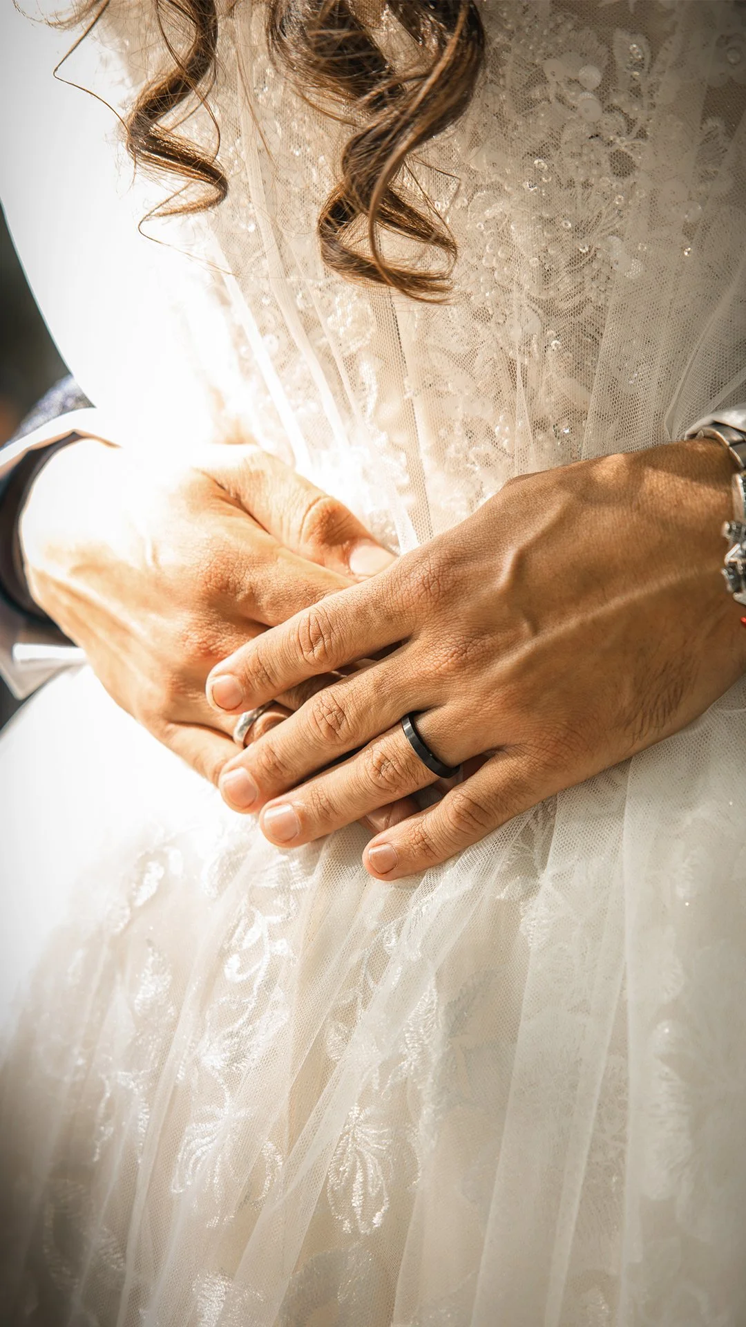 Primeiro plano de mãos entrelaçadas de um casal, com alianças de casamento, em um vestido de noiva com detalhes de renda e lantejoulas, simbolizando um momento de casamento.