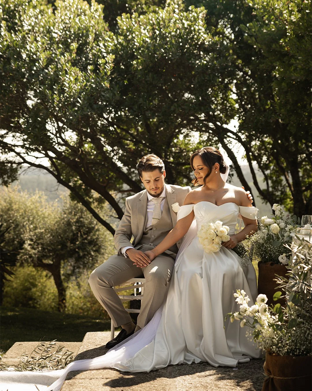 Casal de casamento ao ar livre, com o noivo de terno cinza e a noiva de vestido branco longo, sentado em degraus de pedra rodeados por árvores verdes e flores brancas.