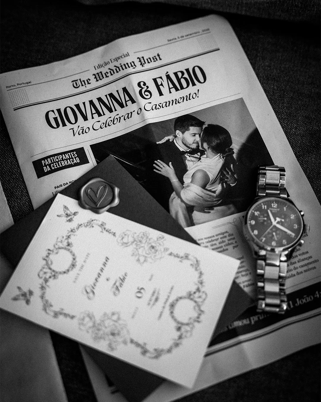 Matéria de jornal com anúncio de casamento de Giovanna e Fábio, uma foto de um casal se beijando, um convite de casamento, um relógio de pulso de metal e um envelope com selo de assinatura de coração.