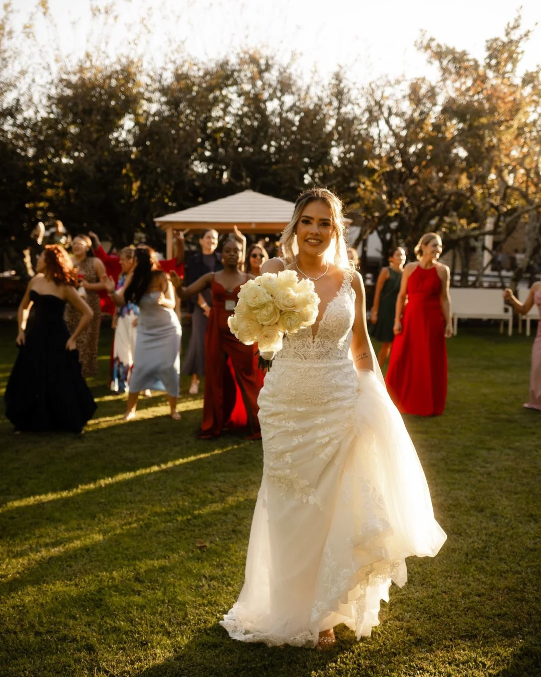 Noivos e convidados celebrando ao ar livre ao pôr do sol, com a noiva em vestido de casamento segurando buquê de flores brancas, cercada por muitas mulheres em vestidos elegantes.