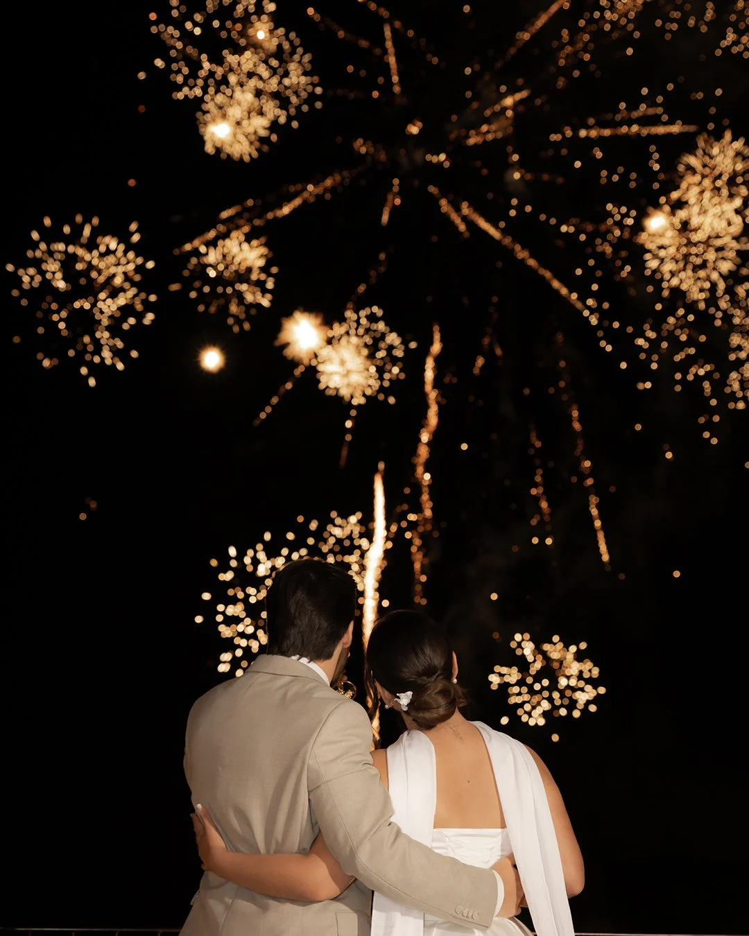 Casal de noivos assistindo fogos de artifício ao fundo em noite escura, com eles de costas, um com terno bege e outro com vestido branco.