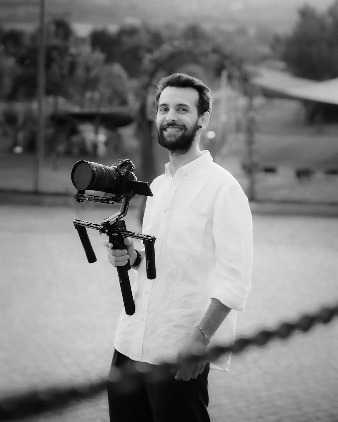 Homem sorridente segurando uma câmera em um estabilizador, ao ar livre, com árvores ao fundo, em preto e branco.