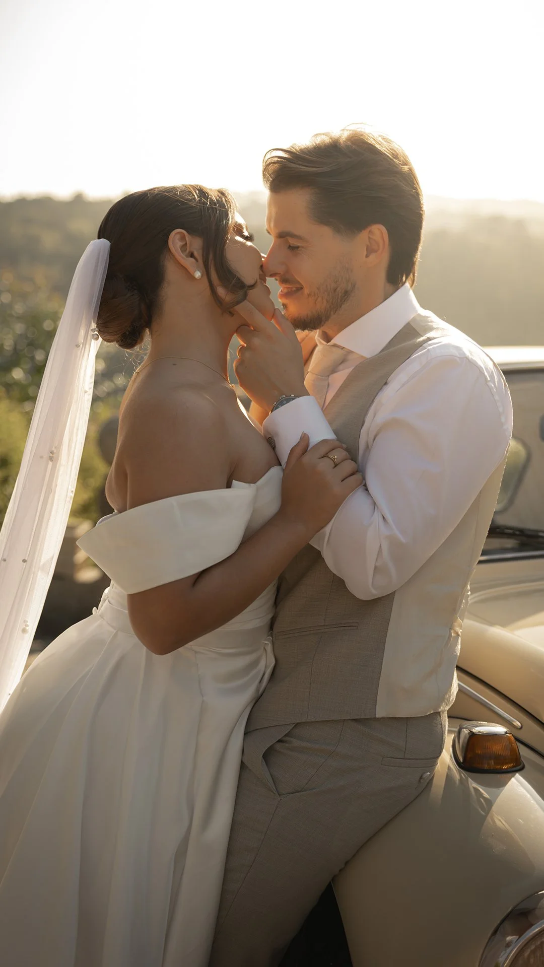 Casal de noivos em momento romântico, com o noivo segurando o rosto da noiva, ambas sorrindo e com os olhos fechados, ao entardecer.