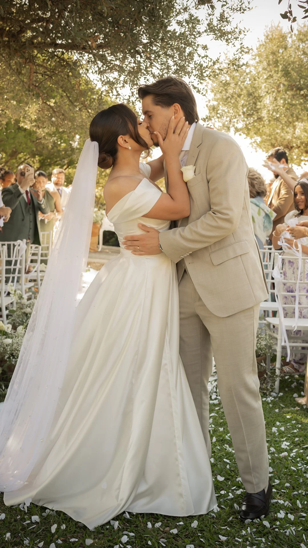 Casal de noivos beijando, ao ar livre, com convidados ao fundo e árvores ao redor.