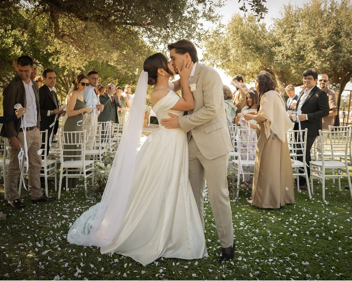 Casal de noivos se beijando durante casamento ao ar livre, com convidados ao fundo e decoração de flores e cadeiras brancas.
