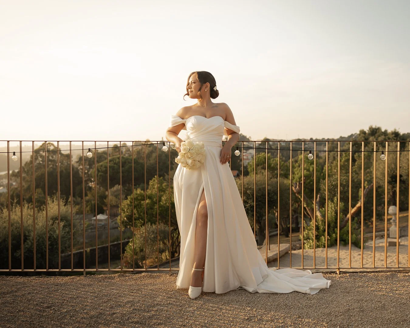 Mulher em vestido de noiva segurando buquê, na varanda ao entardecer, com cenário de árvores ao fundo.