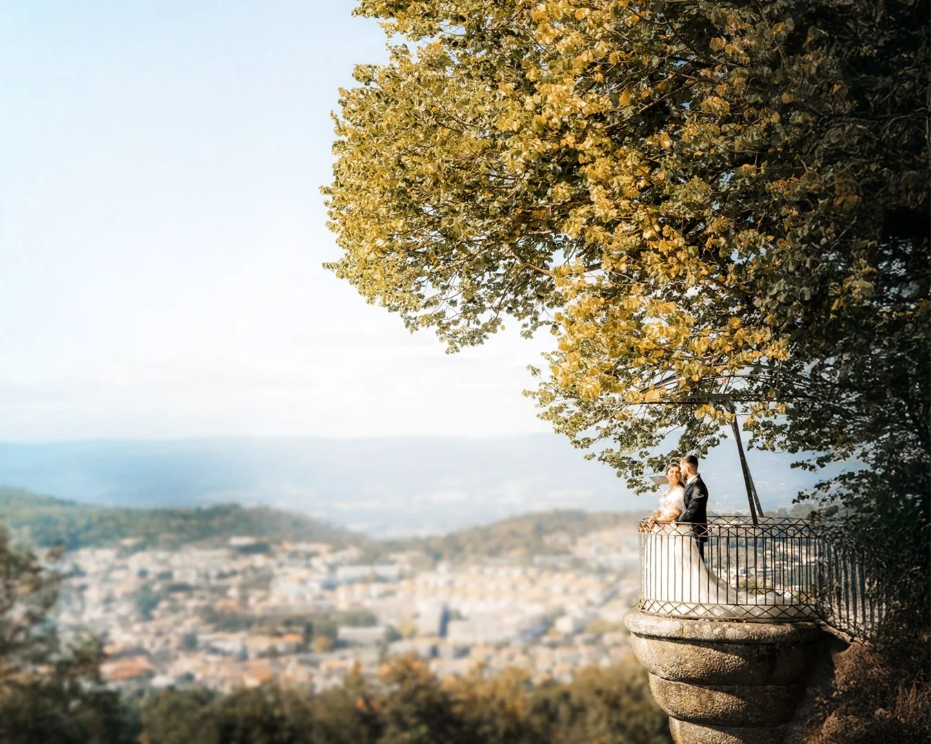 Casal de casamento em um balanço sob uma árvore em uma colina com vista para uma cidade ao longe.