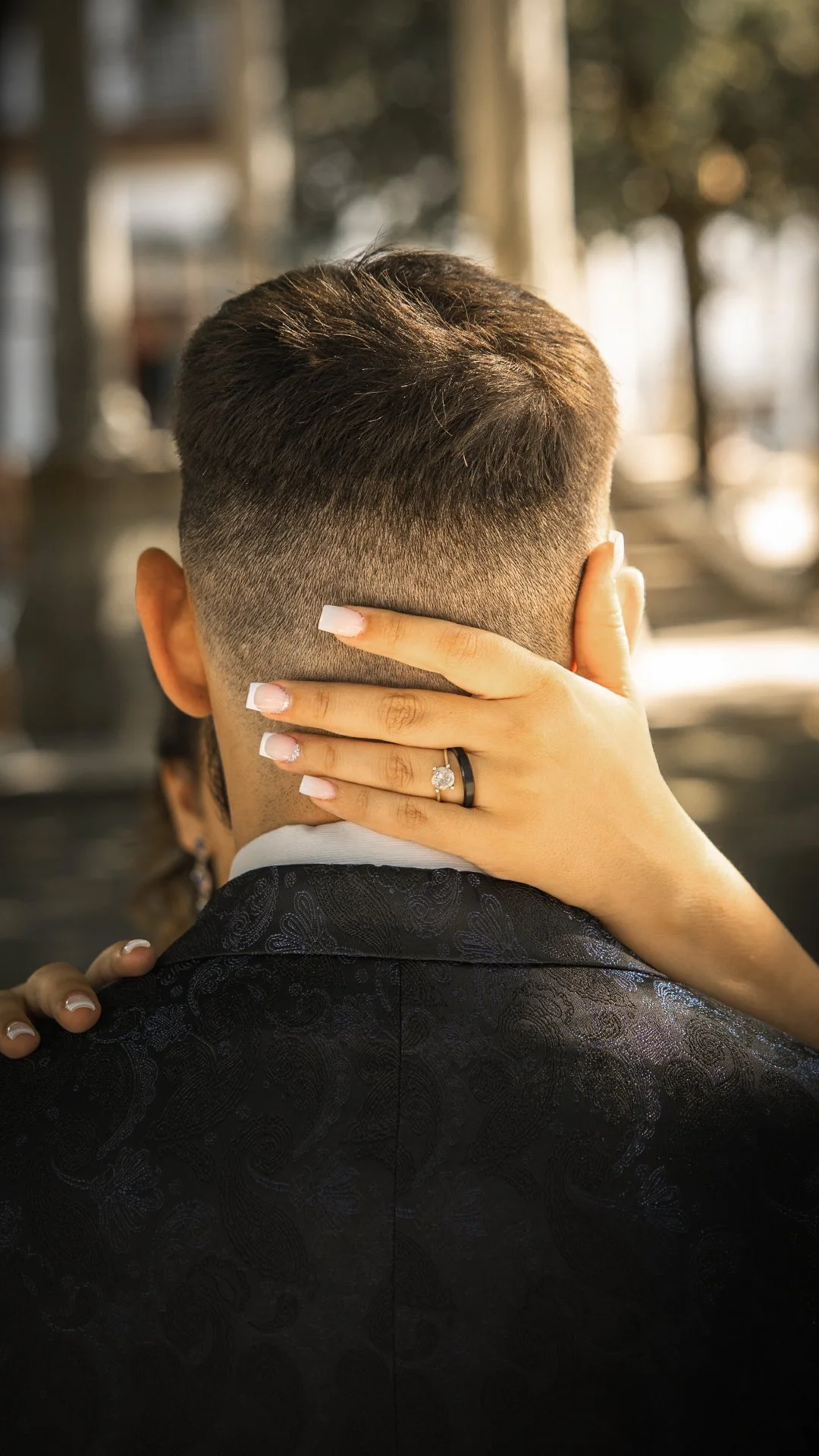 Pessoa com cabelo curto e bem cuidado, usando terno preto com detalhe em padrão, com uma mão apoiada na cabeça de outra pessoa, que está usando uma aliança de noivado ou casamento.