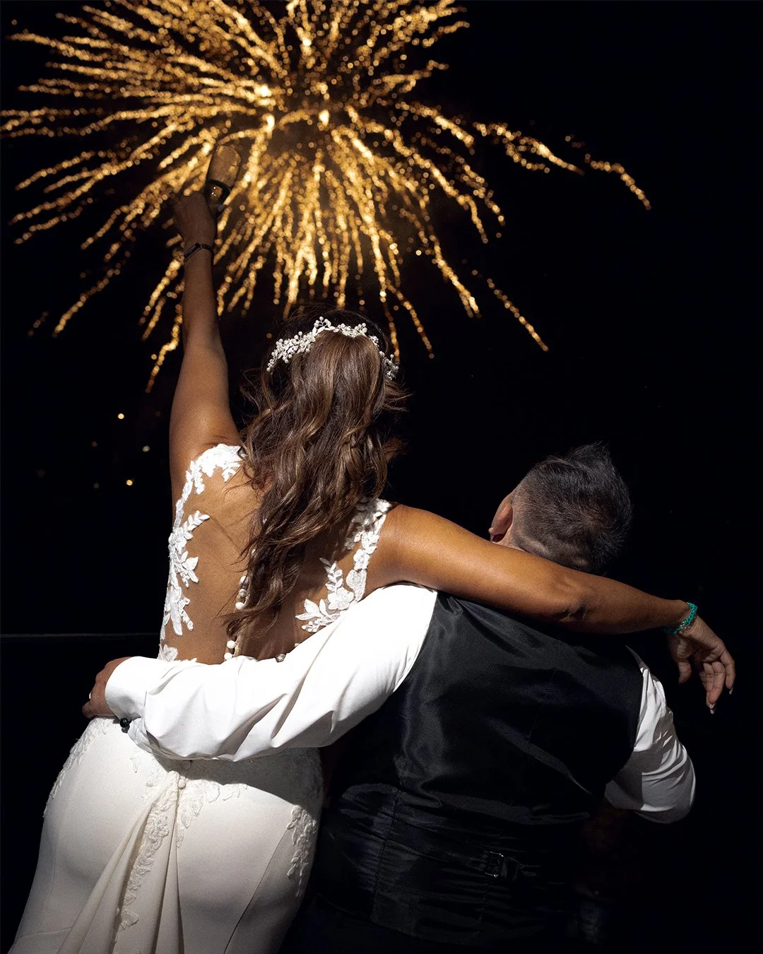 Casal comemora casamento com fogos de artifício ao fundo, ela usando vestido de noiva e ele vestindo terno, noite de celebração.
