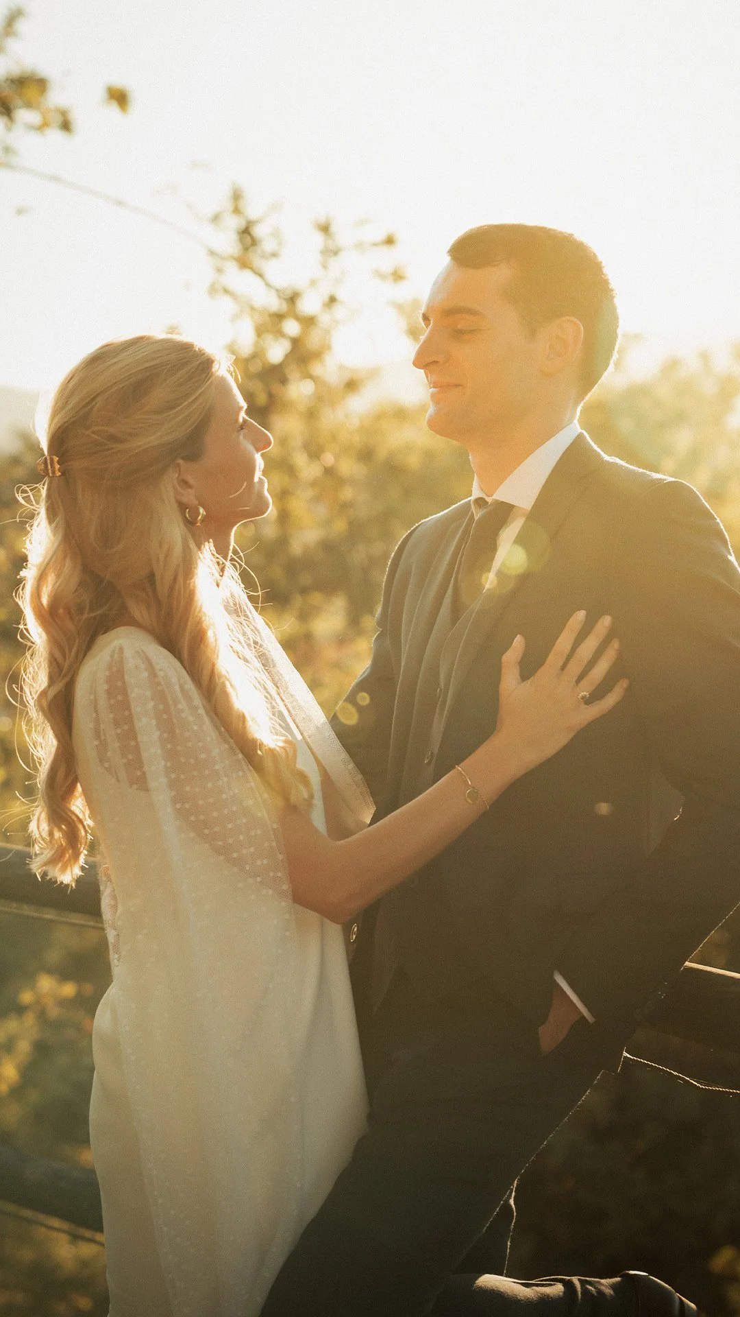 Casal em um casamento ao ar livre, com o homem de terno e a mulher com vestido branco, sorrindo e olhando um para o outro sob luz do pôr do sol.