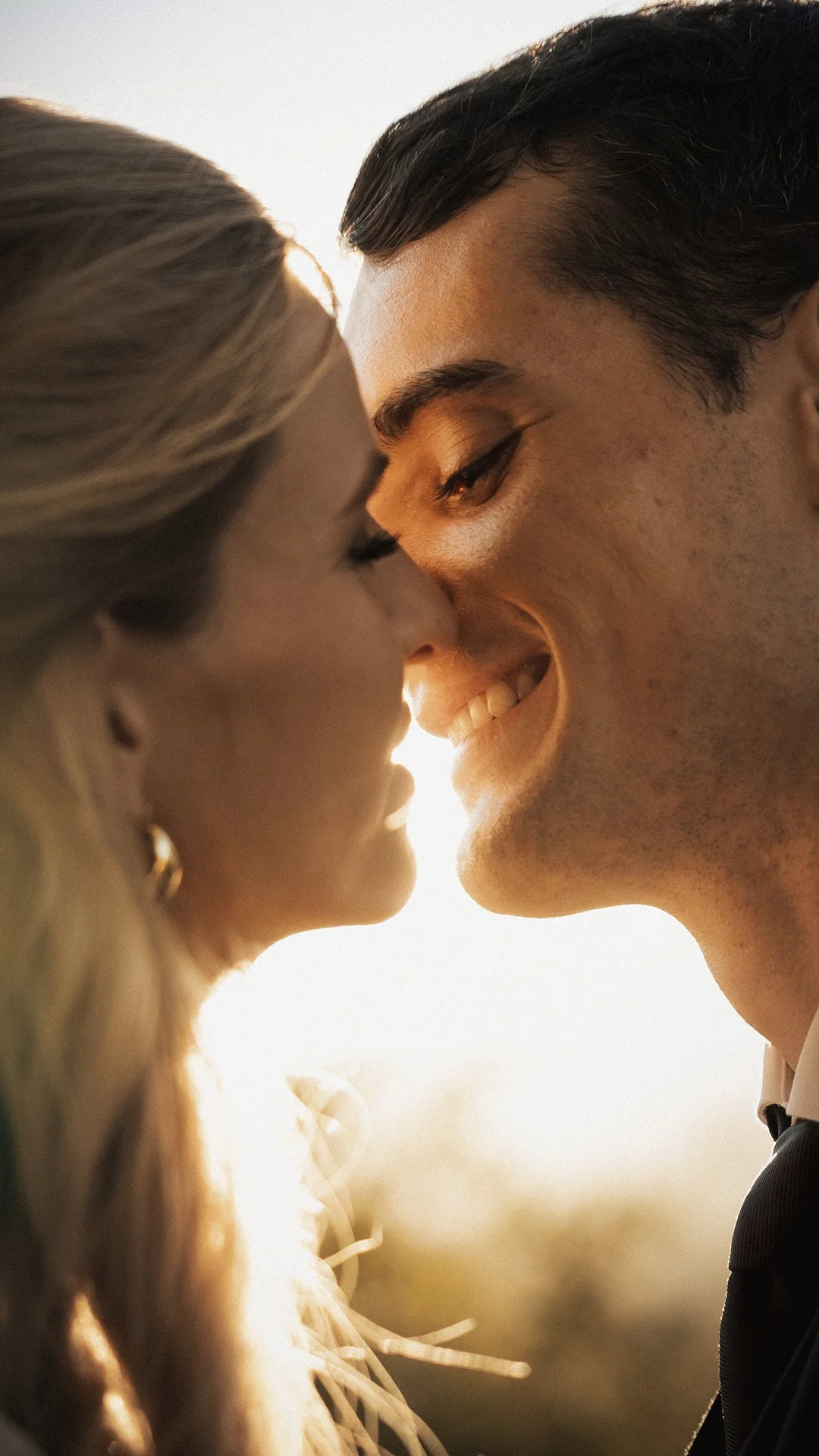 Um casal sorri com o rosto quase tocando, compartilhando um momento de carinho ao entardecer.