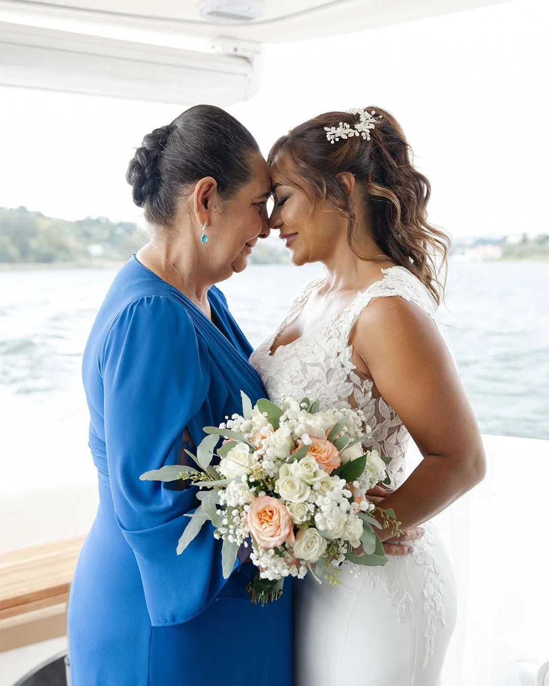 Duas mulheres se abraçando e tocando a testa uma na outra em um cenário de festa na água, uma vestindo vestido de noiva branco e a outra vestido azul, segurando um Buquê de rosas brancas e rosadas.