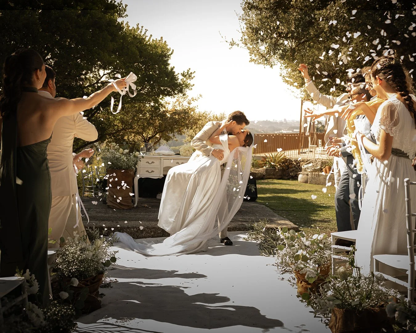Casal de noivos se beijando durante cerimônia de casamento ao ar livre com convidados lançando pétalas de flores e confetes ao entardecer.