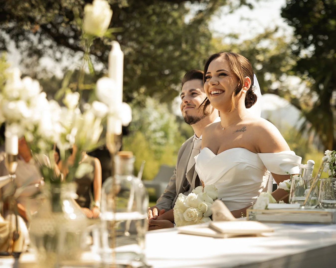 Casal de noivos sentados em uma cerimônia ao ar livre, sorrindo e apreciando o momento, com decoração de flores brancas na mesa e ao fundo árvores verdes.