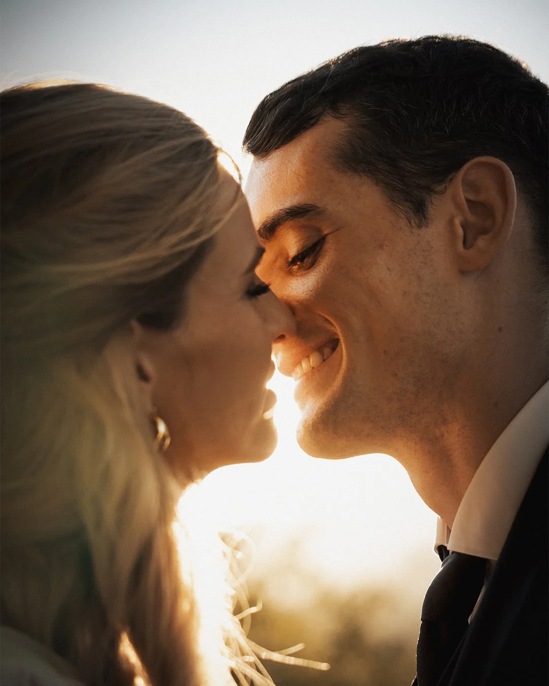 Um casal se aproxima com os rostos quase tocando, sorrindo e trocando um olhar íntimo em um ambiente bem iluminado com luz natural ao entardecer.