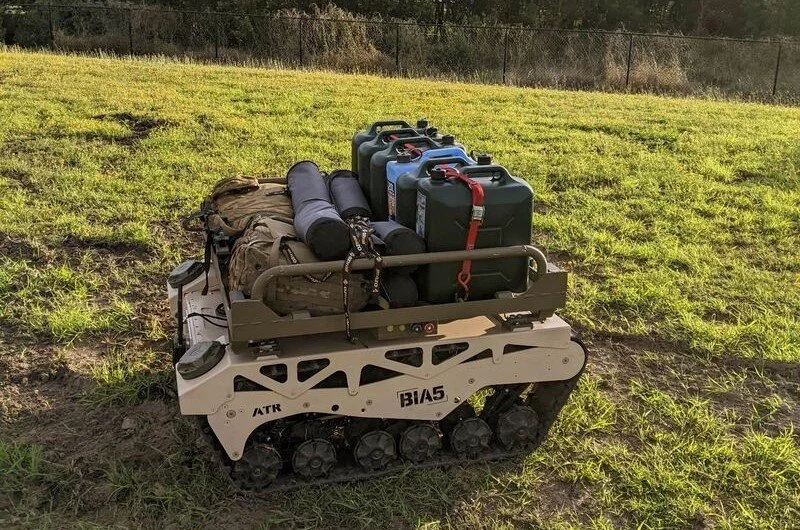 A small tracked robot carrying a backpack, three gas cans, and additional gear on a grassy field.