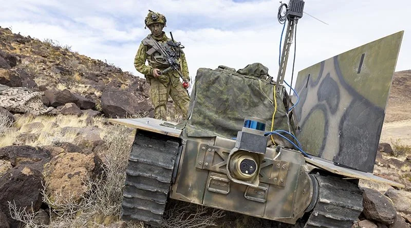 Military robot in desert terrain with soldier in camouflage uniform standing behind it, holding a rifle.