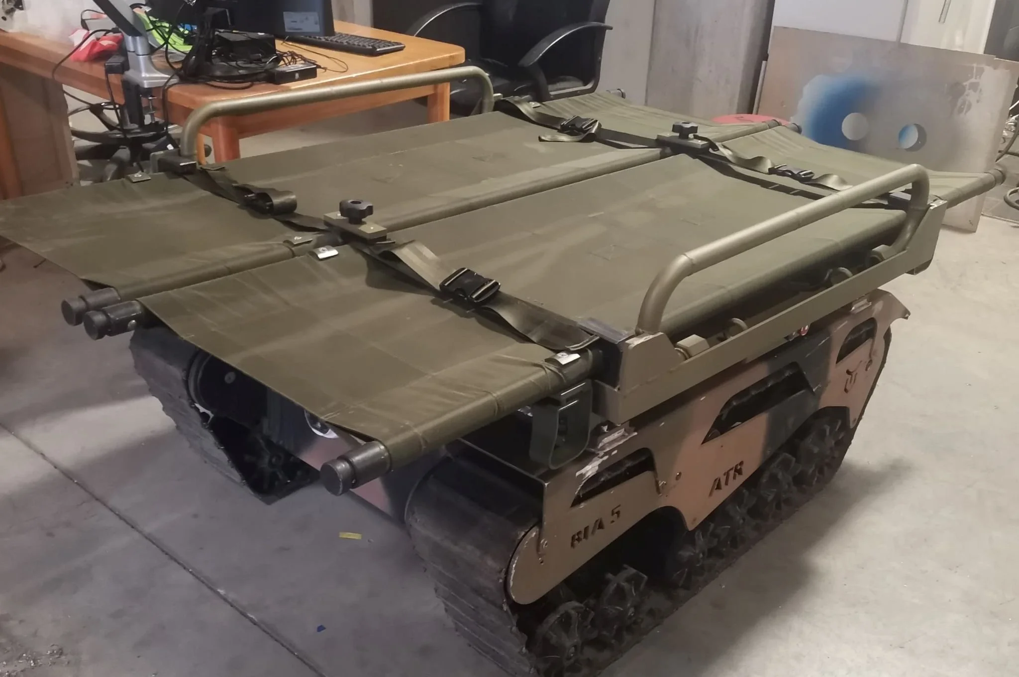 A military-style foldable cot with a green fabric surface and metal frame, placed on top of a storage container in an indoor setting.